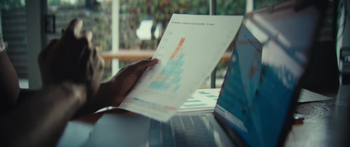A close view of a man holding a document with a bar graph while sitting in front of a laptop at a wooden table in a bright modern home