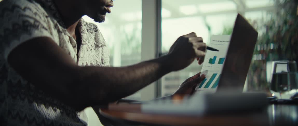 A man sitting at a table while reviewing a document featuring several colorful bar charts he uses a pen to point at specific data points on the page while a laptop remains open in front of him
