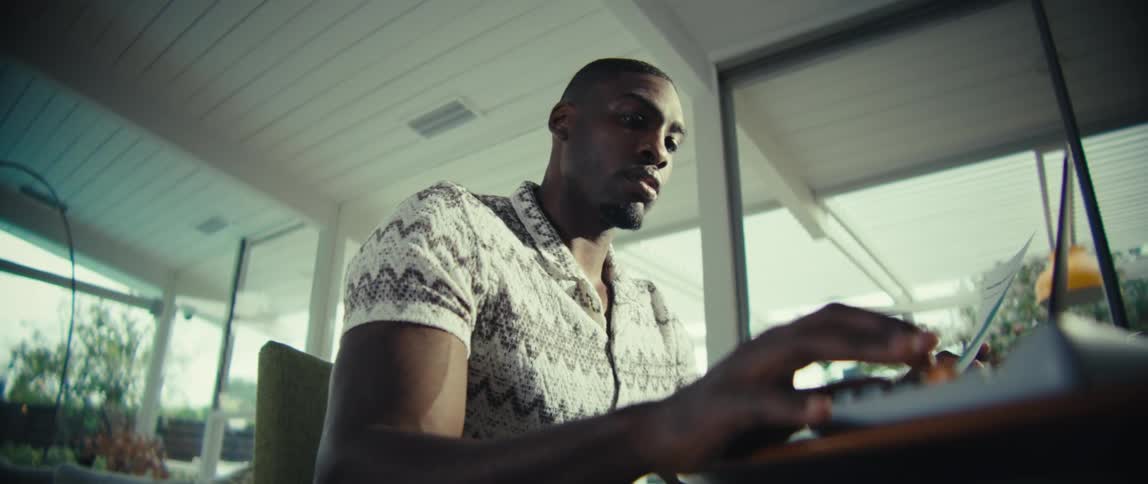 A black man sits at a desk typing on a calculator and reading on a laptop in a modern sunlit room