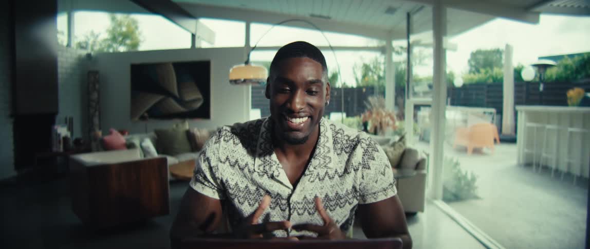 A man wearing a patterned short sleeve shirt sits at a wooden table and smiles while waving at a laptop camera during a video call in a bright modern home