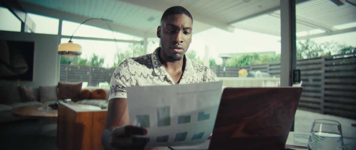 A medium shot of a man sitting at a wooden table in a brightly lit modern room he is focused on reviewing a document with charts and graphs while working on a laptop with a woodgrain finish