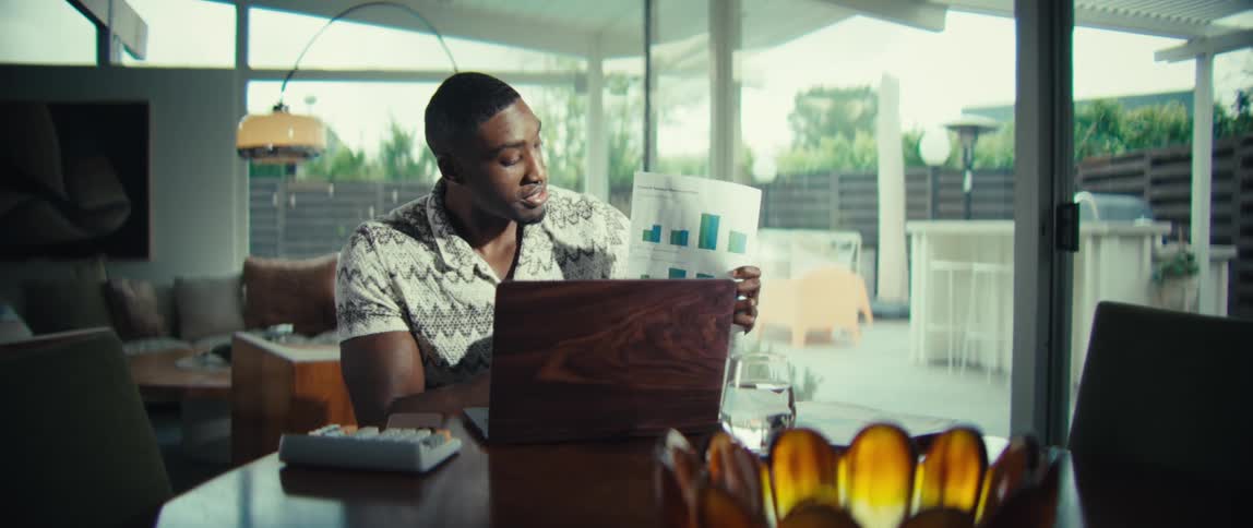 A man wearing a patterned short sleeve shirt sits at a wooden table and holds up a document with several colorful pie charts while speaking during a video call on a laptop in a bright modern home