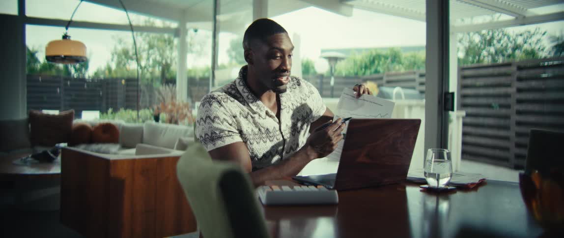 A man wearing a patterned shirt speaks while holding a document with a line graph and pointing at it with a pen while sitting at a dining table with a laptop in a bright modern home