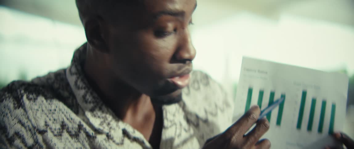 A man wearing a patterned short sleeve shirt sits at a wooden table and holds up a document with green bar charts while pointing and speaking during a video call on a laptop in a bright modern home