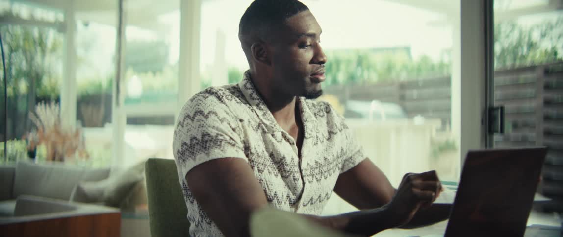 A black man sits at a table and talks during a video call on his laptop while holding up a printed document featuring several bar charts to show data in a bright room