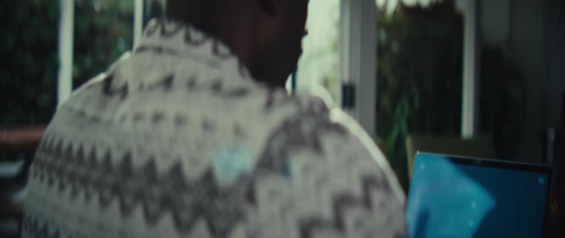 A man wearing a patterned sweater sits at a desk and types on a laptop the camera moves from behind his shoulder to reveal the back of his head as light filters in from a nearby window