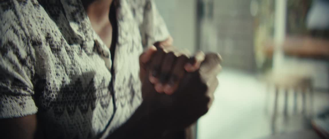 A smiling black man sits at a desk looking at a laptop rubbing his hands together in a sunlit room