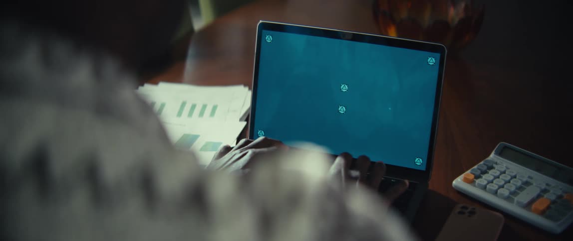 A person is seen from an overtheshoulder perspective while they type on a laptop at a wooden deskthe laptop screen is a solid blue color with several tracking markers and it is surrounded by items like printed charts and a calculator
