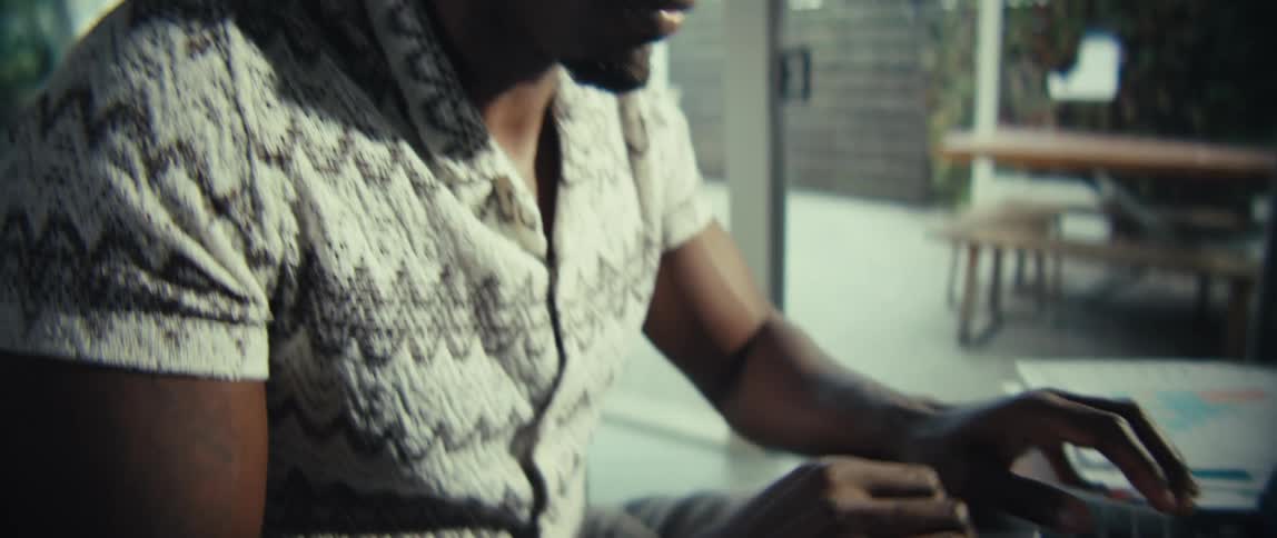A black man sits typing on a laptop in deep thought at a cluttered table in a sunlit modern room