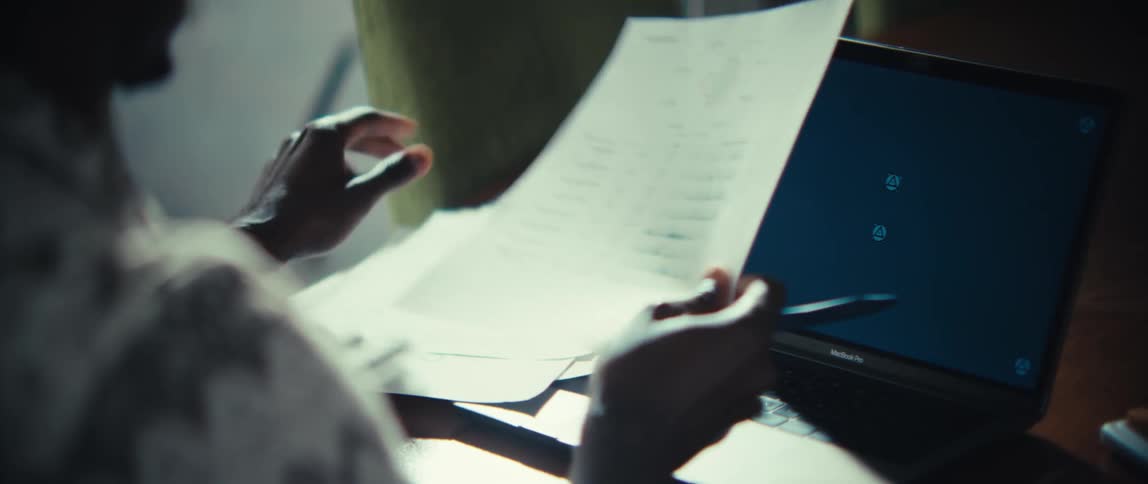 Man sitting at a table in front of a laptop computer holding a pen as he looks through documents