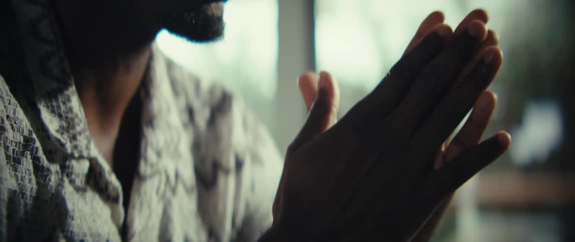 A close view of a man with a beard looking forward with a serious expression and then clasping his hands together before typing on a laptop keyboard in a bright room