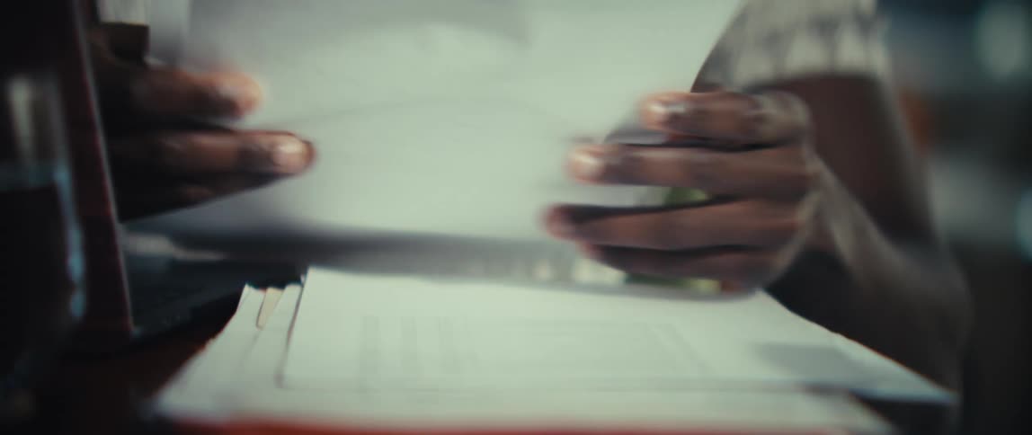 A man wearing a patterned shirt looks through a stack of papers with charts and data while sitting at a table with a laptop in a bright room