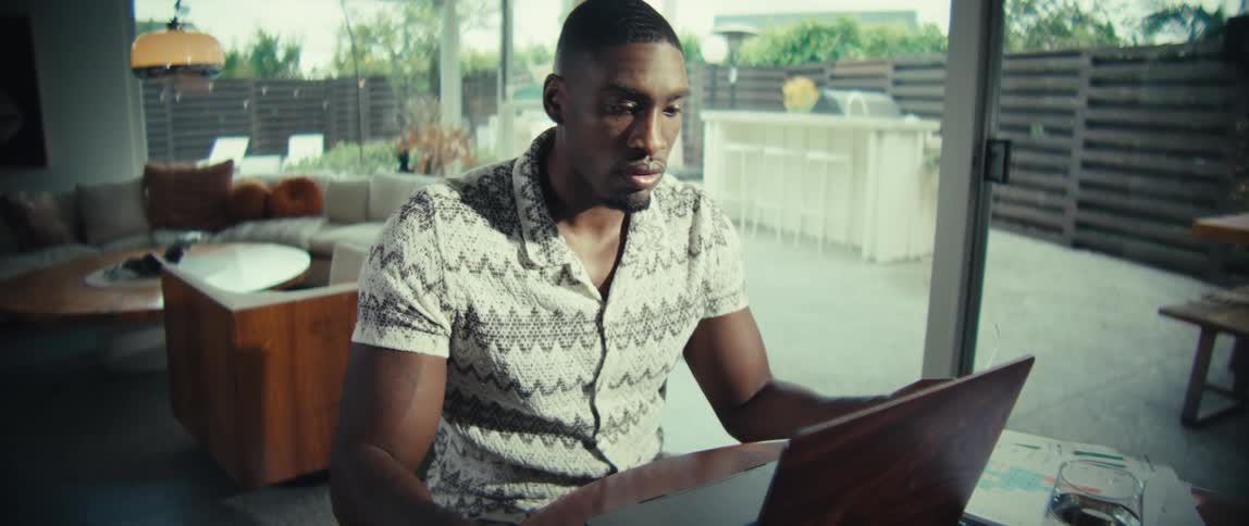 A man wearing a patterned short sleeve shirt sits at a wooden table and looks at his laptop screen with a serious and focused expression while working in a bright modern home