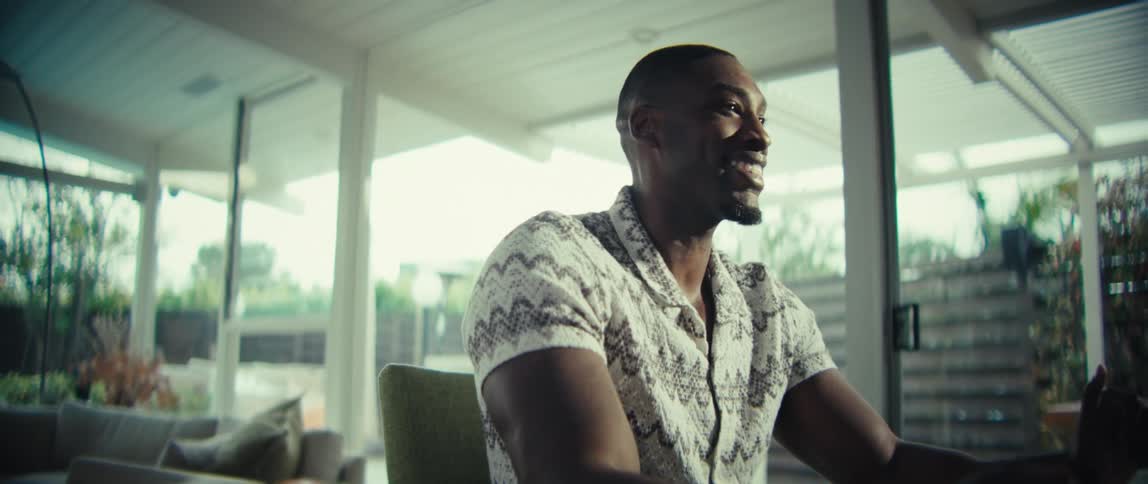 A man seated at a table with his laptop sits back and smiles