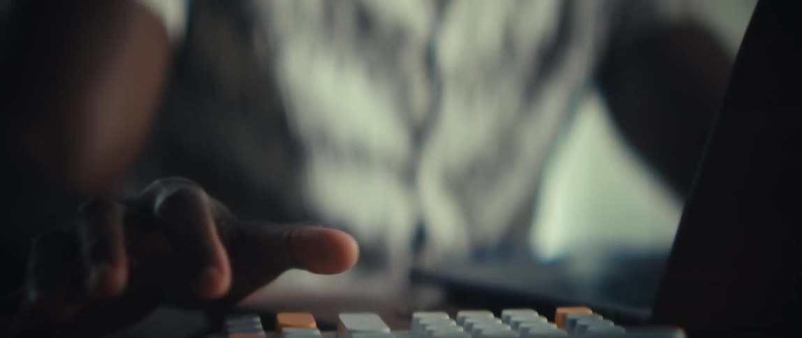 A black man sits at a table in a modern room typing on a calculator and looking at a tablet on the table