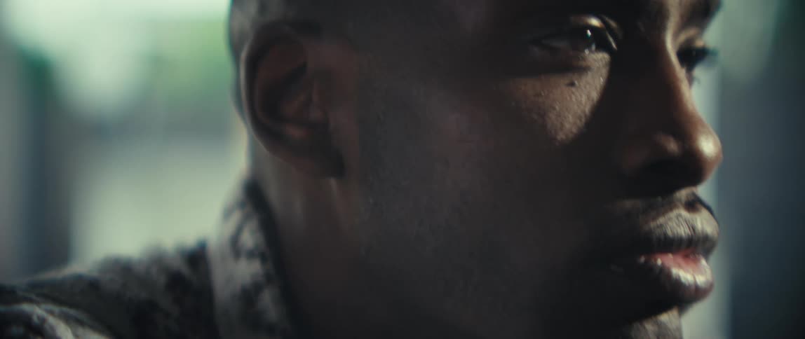 A close view shows the face of a man looking away with a serious expression before the camera moves to show his hands clasped together