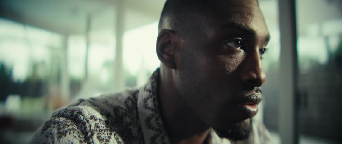 A black man sits clasping his hands in sadness before typing on a laptop and calculator in a sunlit room