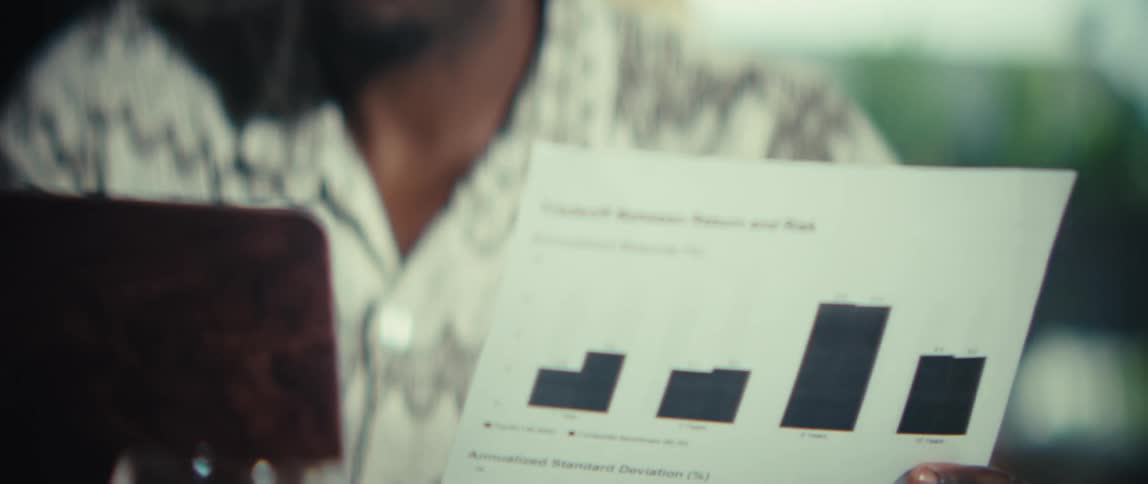 A man wearing a patterned short sleeve shirt sits at a wooden table and smiles while looking at a document with bar charts and then at a laptop during a bright day in a modern home