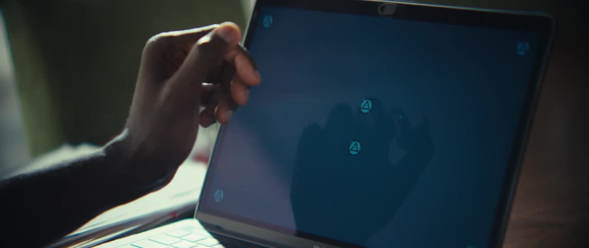 A black man is shown typing on a laptop and a calculator while sitting at a desk in a dimly lit room