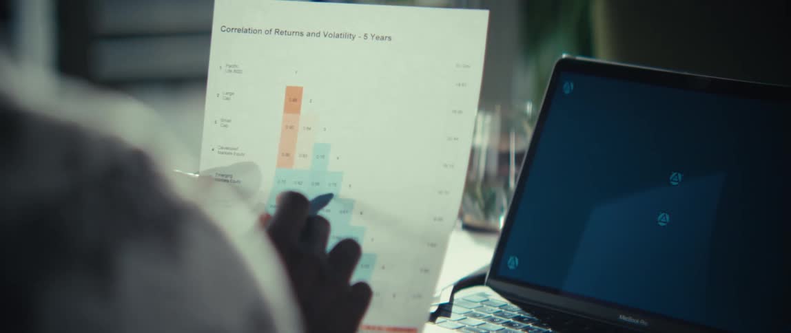 A man wearing a patterned shirt reviews a financial document with a pen and then uses the pen to point at a laptop screen in a close view