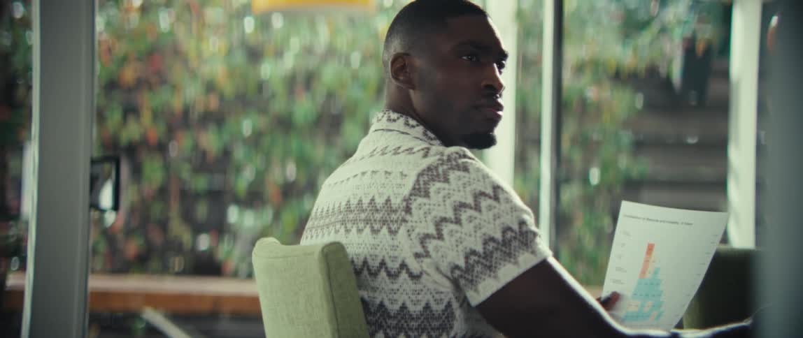 A man wearing a patterned shirt holds a document with data and look over his shoulder with a serious expression in a bright room with a large window