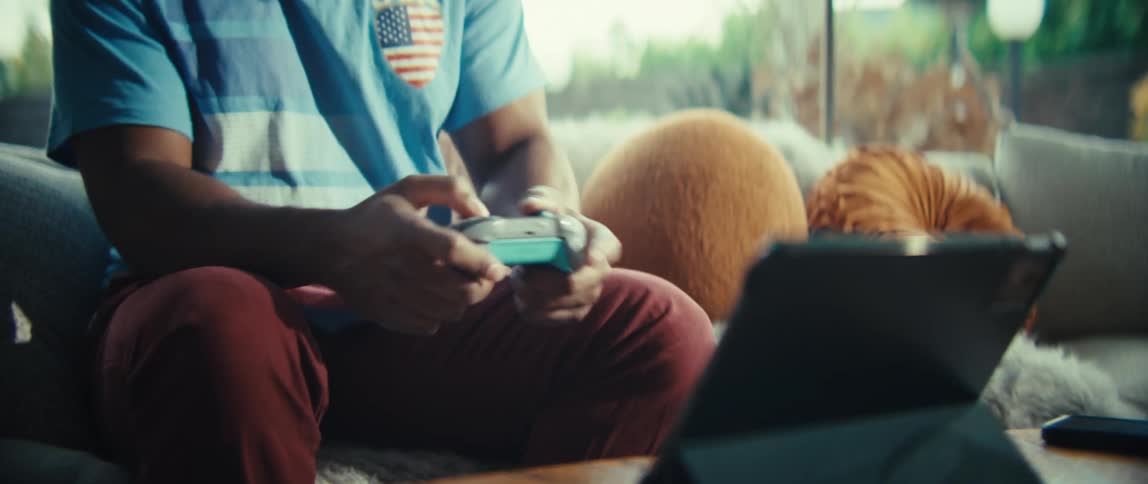 A close view shows the hands of a man as he presses buttons on a blue and grey game controller while sitting on a sofa in a bright room