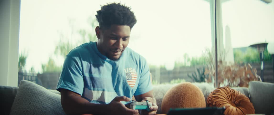 A man wearing a blue shirt smiles and looks at a screen while using a game controller while sitting on a sofa in a bright living room