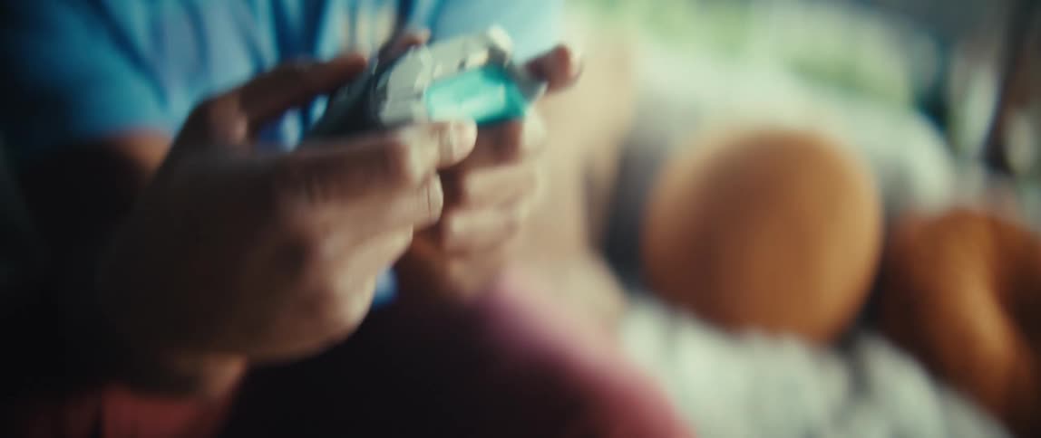 An excited black man sits on a couch in a sunlit room using a game remote control