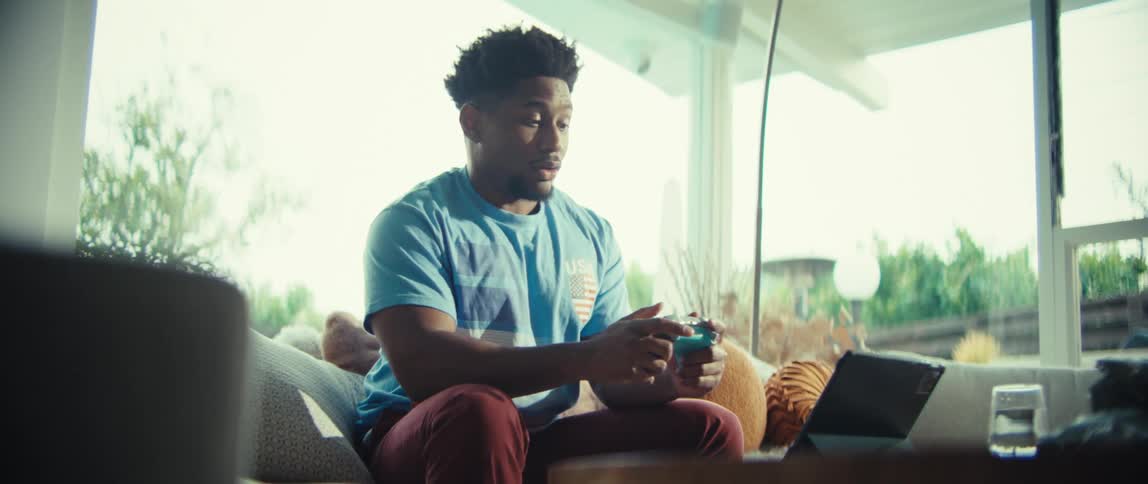 A man wearing a blue shirt smiles and speaks while holding a game controller and looking at a tablet on a table while sitting on a sofa in a bright living room