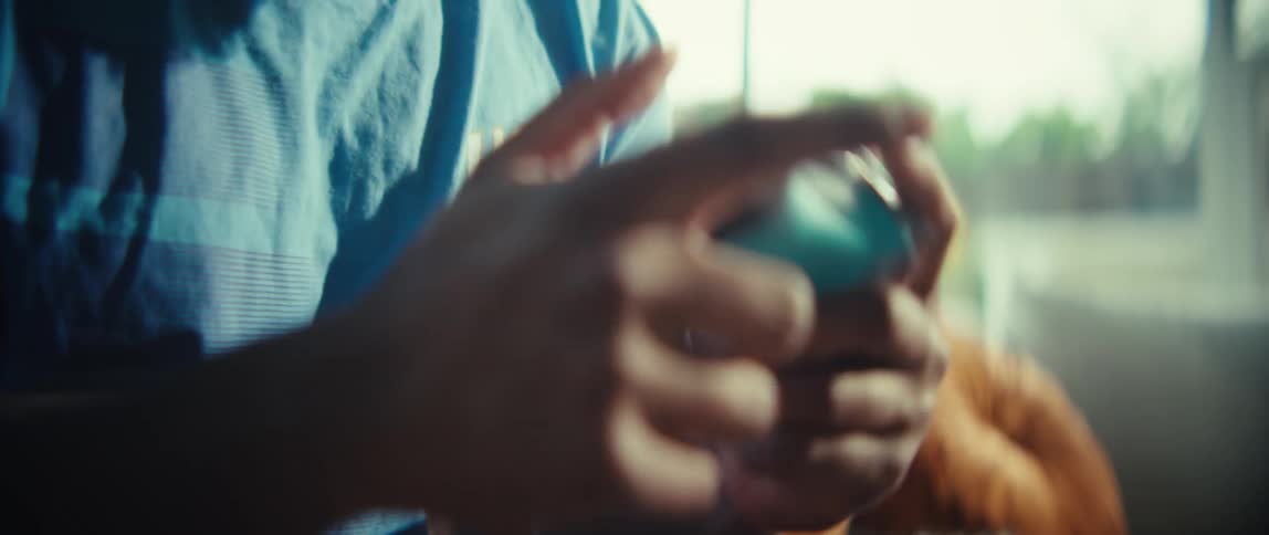 A close view shows the hands of a man as he presses buttons on a blue and grey game controller while sitting in a bright room