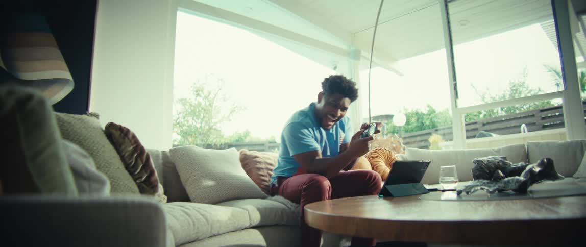 Young man sitting on a couch holding a video game controller as he plays a game on a tablet computer and laughs in excitement