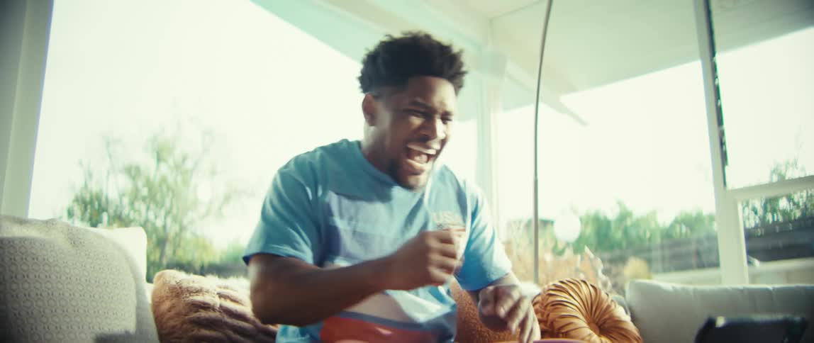 A man wearing a blue shirt with a usa graphic sits on a couch and watches a tablet before cheering and clapping with a joyful expression in a bright living room