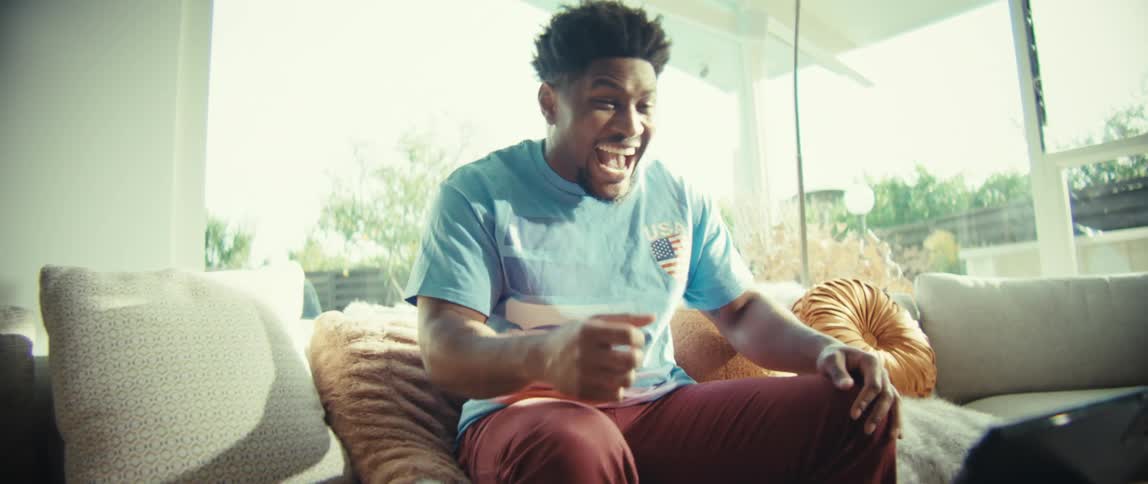 A man sitting on a couch excitedly pumps his fists while reacting to something on a tablet in a bright modern living room