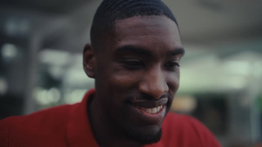 A young black man with a short haircut wears a red polo shirt while smiling and typing on a laptop computer at a wooden table in an outdoor setting