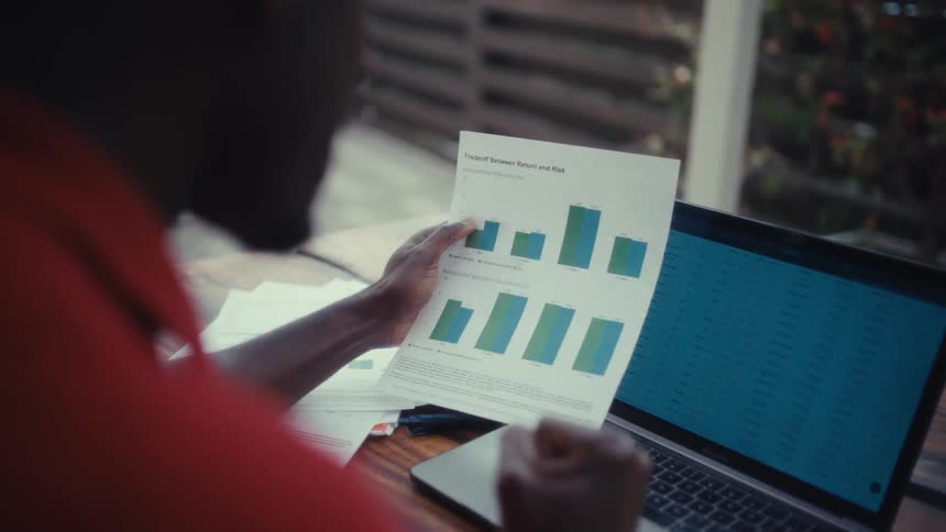 Person holds a sheet with a bar chart beside an open laptop on a table