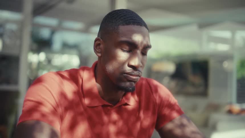 A young black man wearing a red tshirt sits outdoors at midday resting his hands on his laptop while looking sad and deep in thought