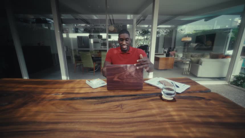 Man closes a laptop on a wooden table in a space with glass doors in the background