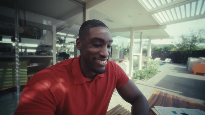 A man in a red polo shirt sits at a long wooden table on a sunny patio focused on typing on his laptop he eventually pauses his work to look at the screen and shares a warm cheerful smile