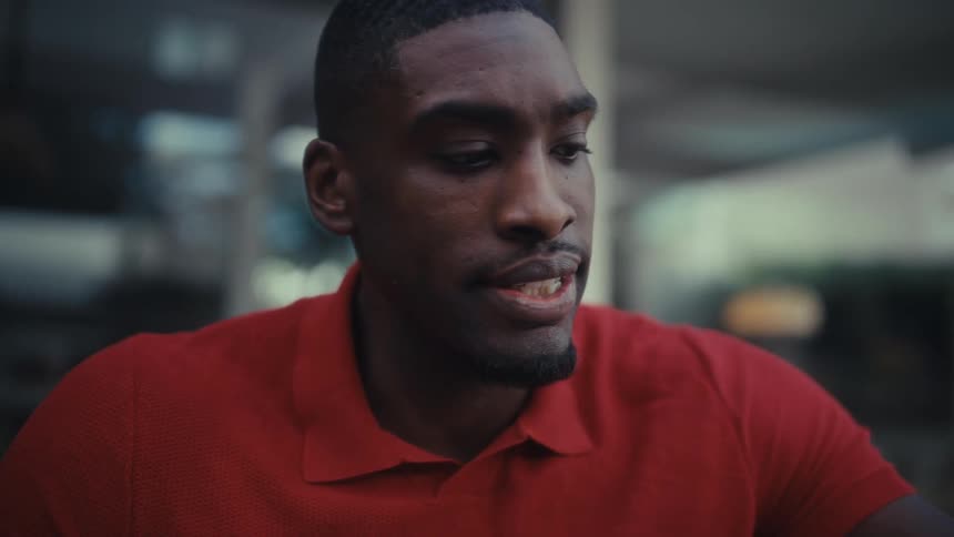 A young black man wearing a red tshirt sits outdoors at midday takes a sip from a glass of water and then begins typing on the laptop in front of him