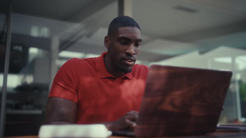 A smiling man in a vibrant red polo shirt sits at a polished wooden desk in a modern sundrenched office his fingers moving rhythmically across the laptop keyboard as he focuses intently on his work against a backdrop of large windows