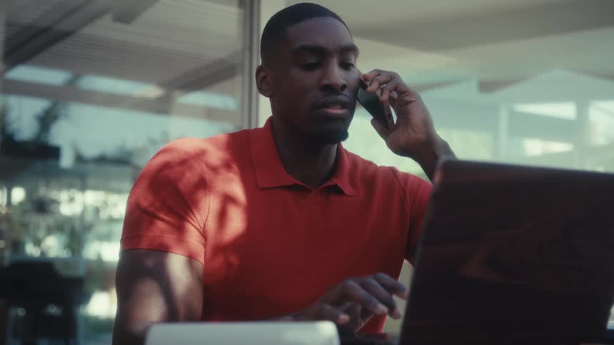 A young man wearing a red tshirt sits outdoors at midday typing on a laptop before picking up his phone to answer a call