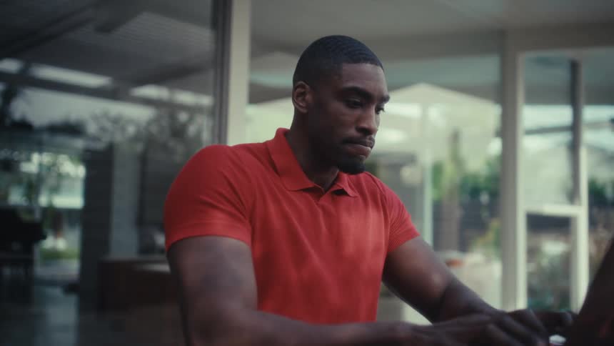 A black man is sitting outdoors on a patio at a wood table working on a laptop and drinking water