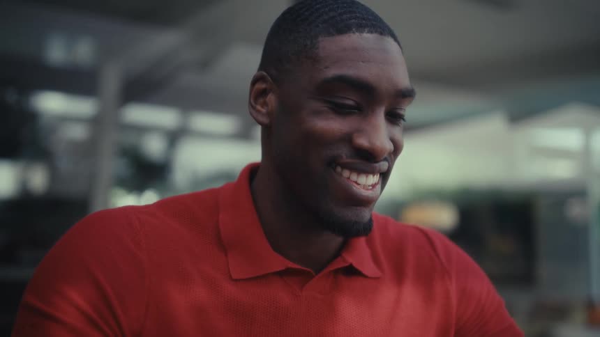 A smiling black man in a red polo shirt sits at an outdoor patio while looking to a paper sheet