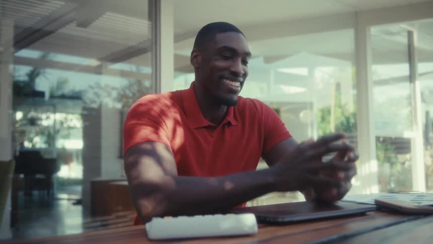 A man wearing a red polo shirt types on a laptop at a wooden table then closes the screen and smiles while looking away in a bright room with large glass windows