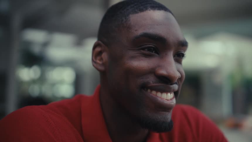 A man typing on a laptop and smiling while looking toward the screen he is wearing a red polo shirt and working at a wooden table that also holds a white keyboard
