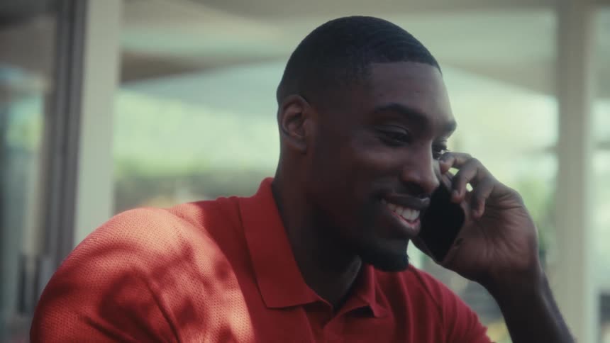 A man wearing a red polo shirt speaks on a mobile phone and types on a laptop while sitting at a wooden table and laughing in a bright room