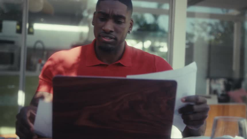 A man in an orange polo shirt sits at a wooden desk focused on reviewing several documents and a laptop he looks between the papers and the screen with a serious concentrated expression as he organizes his work