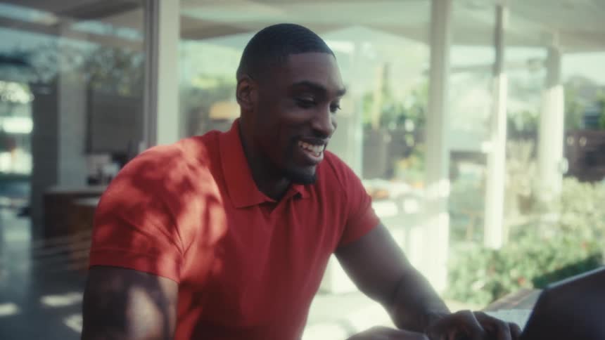 A smiling black man in an orange polo shirt sits at a wooden table on a sunny patio while typing on his laptop