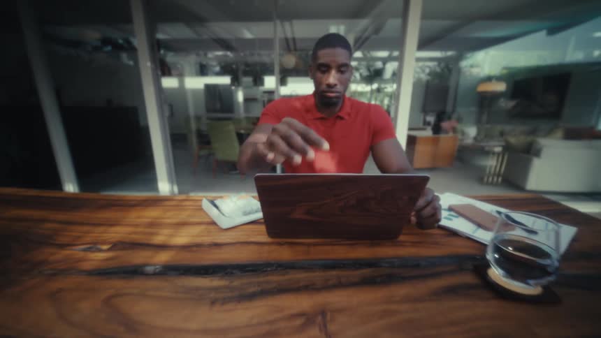 A man wearing a red polo shirt opens the lid of a wood grain laptop and begins to type while sitting at a large wooden table in a bright modern home