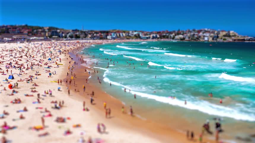 A vibrant shoreline is packed with people sunbathing on the sand and swimming in the turquoise wavesthe footage uses a tiltshift effect that makes the distant buildings and coastal crowds look like a miniature model